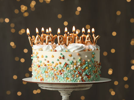 50th Birthday cake with lit HAPPY BIRTHDAY candles, white frosting and pastel sprinkles on a stand against warm lights.