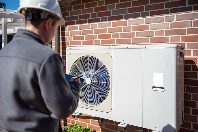 Technician inspecting a white air source heat pump outdoor unit against a brick wall