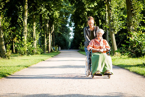 Care worker pushing an elderly resident in a wheelchair along a tree-lined outdoor path