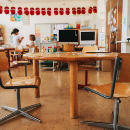 Sunlit classroom with round wooden table and empty chairs in foreground; children work in back under red paper lanterns.