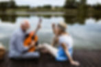 A newly retired senior couple sits on pier playing guitar near a lake