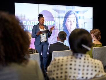 Dark colored woman in a blue business outfit speaking to a group of leaders