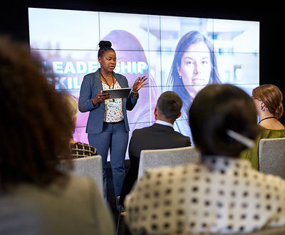A brown-skinned, black executive woman wearing a grey suit stands on stage in front of a well-lit Leadership presentation screen, talking to an audience of corporate employees
