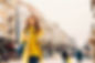 A smiling woman in a yellow coat walks down a sunlit city street