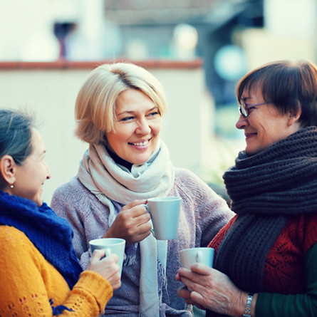 Three women in scarves smiling and holding mugs outside on patio