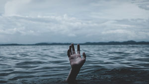 A single hand reaches up from dark, choppy open water beneath a stormy sky, suggesting a person struggling and overwhelmed, as if drowning in deep, threatening conditions.