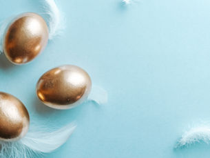 Three gold eggs with white feathers on a light blue background. The scene is minimalist and serene.