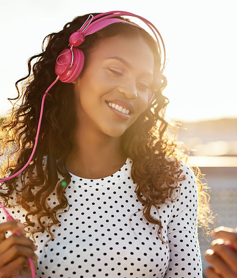 A woman with curly brown hair listen to music on pink headphones outside in the sun