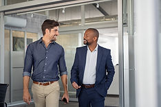 Dois homens caminhando dentro da empresa,conversando e sorrindo. O homem lado esquerdo, é branco, cabelos curtos, preto e liso. Está usando uma camisa azul marinho e uma calça na cor caramelo e cinto na cor marrom.r
