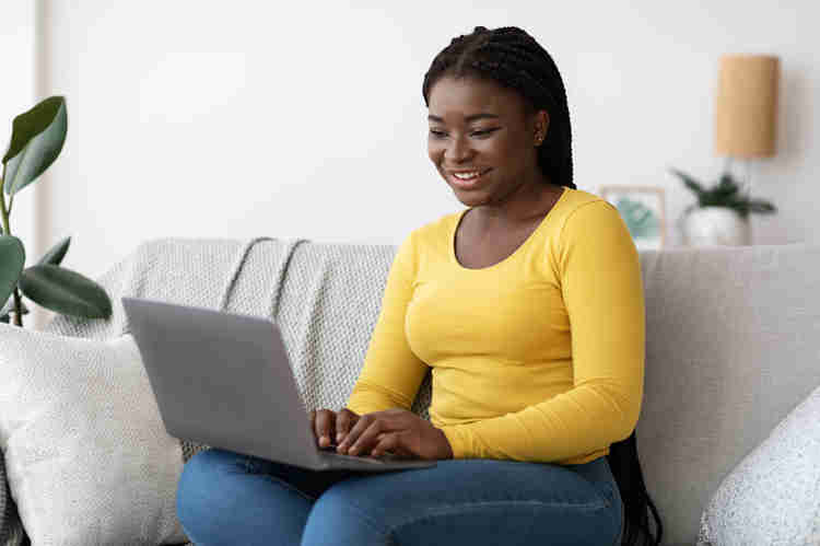 This is a picture of a Black woman sitting on a gray couch. She's wearing jeans and a long-sleeved yellow shirt. On her lap is an open laptop.