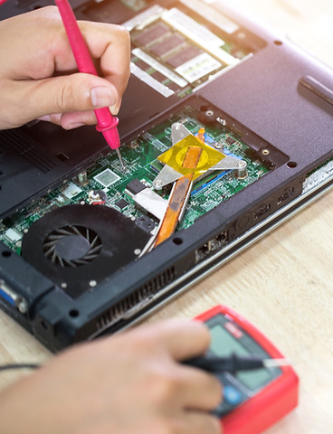 A person repairs a laptop on a wooden table, using a multimeter.