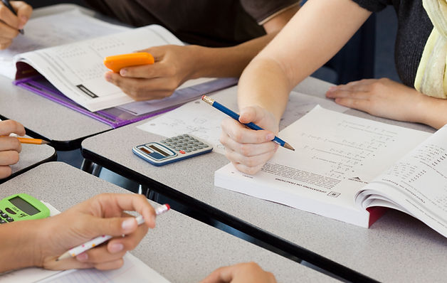 foto das mãos de 4 alunos escrevendo em cadernos e livros , usando lápis nas mãos e calculadoras sobre a mesa