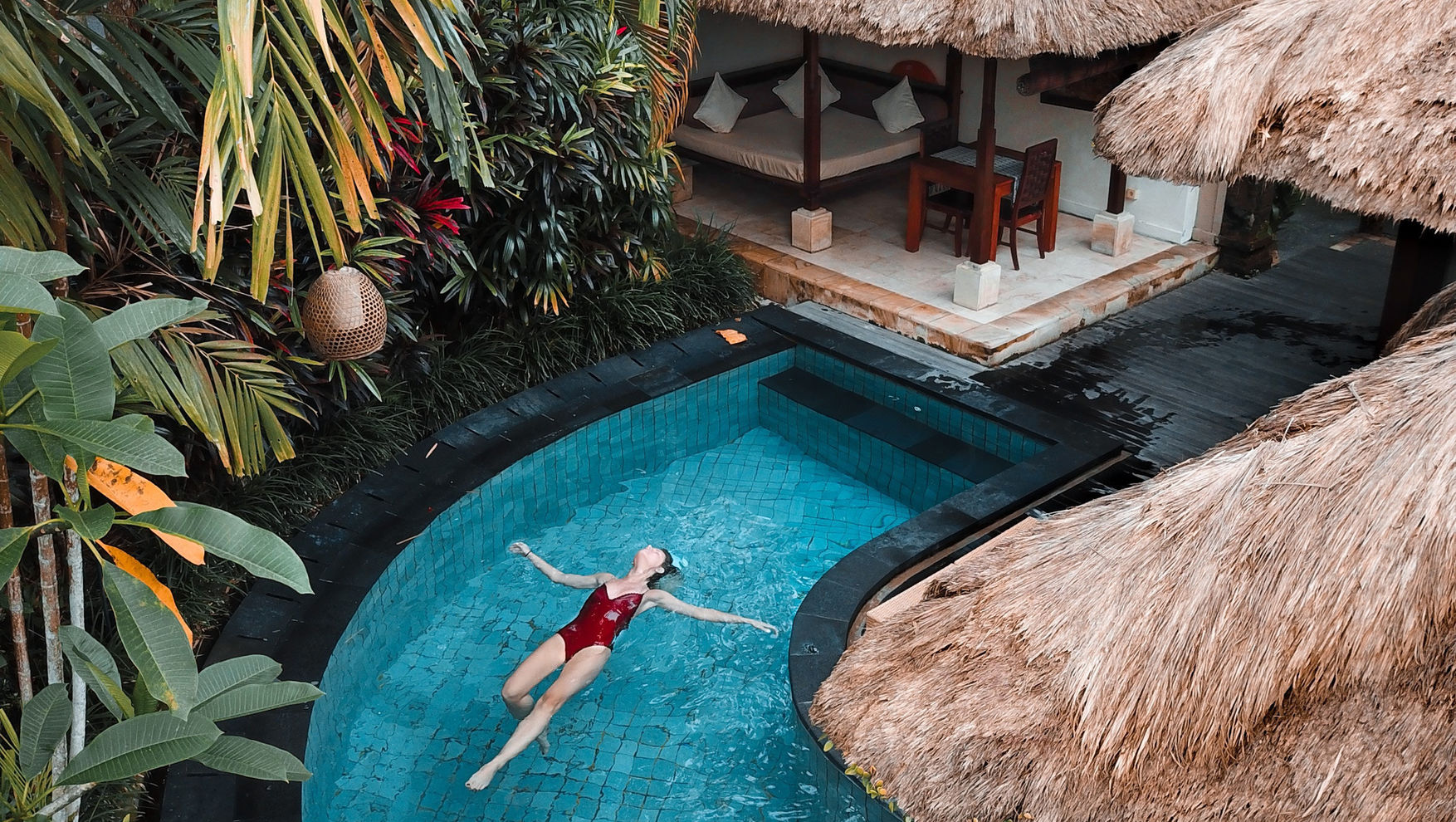 Woman in a red swimsuit floats in a blue-tiled pool surrounded by lush greenery and tropical huts with thatched roofs. Relaxed mood.