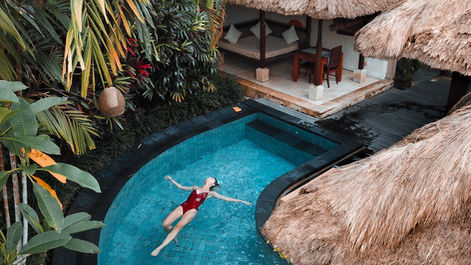 Woman in a red swimsuit floats in a blue-tiled pool surrounded by lush greenery and tropical huts with thatched roofs. Relaxed mood.