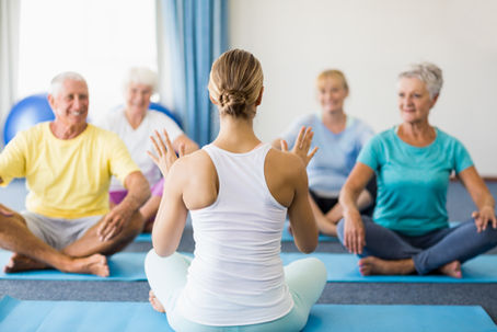 A group of older adults sit cross-legged on yoga mats, guided by an instructor. The setting is a bright room with blue curtains, creating a calm mood.