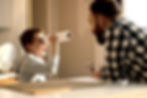 child looking through a tube while smiling and wearing a gray hoodt. Tan skinned person with a beard facing the child and wearing a black and white checkered long sleeve shit. they appear to be in a kitchen. 