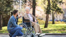 A man kneels beside a smiling elderly woman in a wheelchair, outdoors in a park. Green trees and sunlight create a peaceful setting.