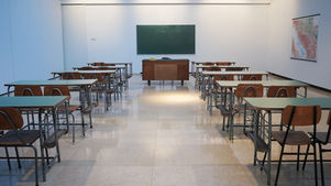 Empty classroom with wooden desks, green chalkboard, and a teacher's desk. A map hangs on the wall. Bright lighting, calm atmosphere.