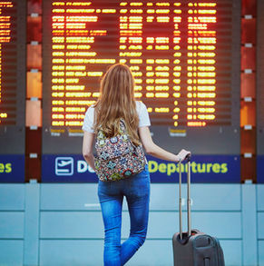 Woman in jeans with patterned backpack looks at an airport departures board. Bright yellow-orange text displays flights. Travel mood. travel insurance ביטוח נסיעות לחו"ל