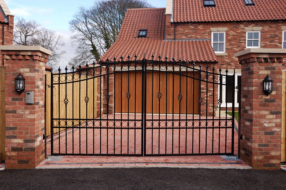 Ornate black iron gate in front of a brick driveway leading to a large house with a red-tiled roof and wooden garage door