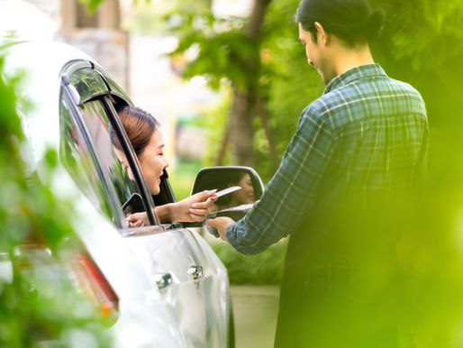Man hands a card to a smiling woman in a white car, viewed through green leaves on a sunny driveway.