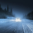Car drives on a snowy forest road at night, headlights illuminating the icy path. Tall, dark trees line the route, creating a moody scene.