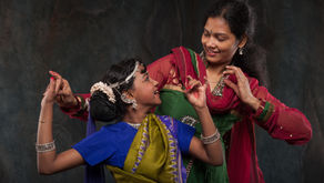 Two women in colorful traditional dress dance together, smiling and looking at each other against a dark studio backdrop.
