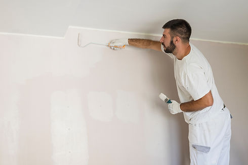 A man in white overalls is painting a light pink wall with a roller