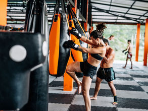 A woman kicks an orange punching bag in a gym with others training nearby. The mood is focused, the space has orange accents and greenery.