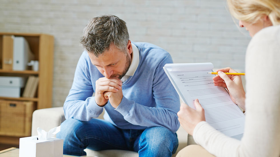 Erkek düşünceli, oturma odasında oturuyor. Kadın not alıyor, form dolduruyor. Ortam sakin, masada bir kutu ve su bardağı var.