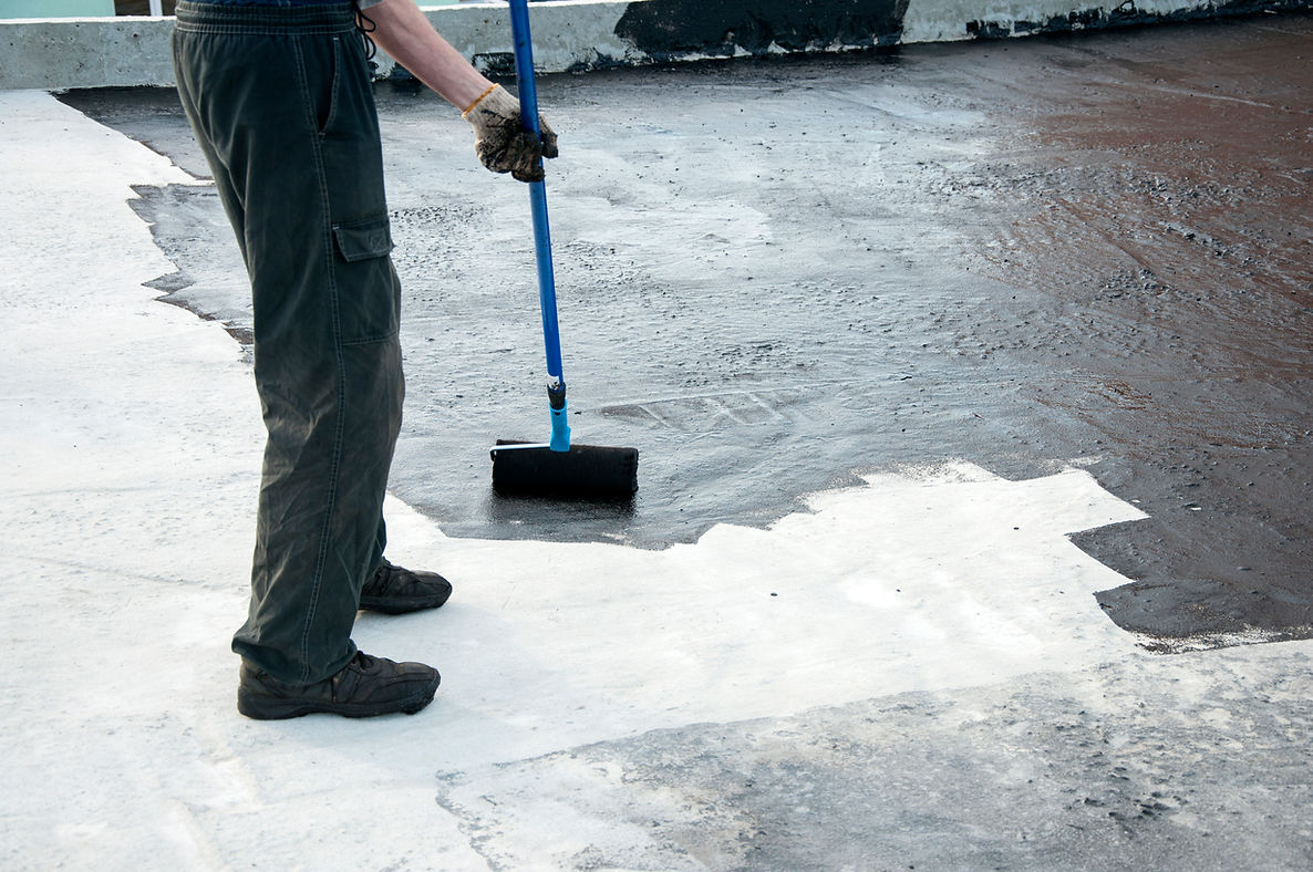 Roofer applying black bitumen primer to a flat concrete surface with a long-handle roller brush