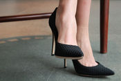 Nahaufnahme: Beine in schwarzen High Heels, ein Fuß angehoben, neben einem Stuhl auf grauem Boden.