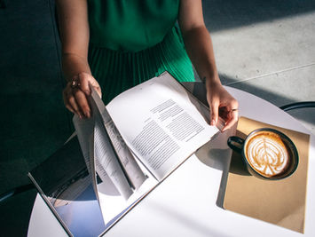 A top-down, editorial-style photograph of a student in a green dress at a white table, hands physically turning the page of a large open book. To the right sits a fresh latte with foam art and a tan notebook. The scene is illuminated by bright, natural sunlight, emphasizing a moment of deep, offline processing within a remote learning lifestyle
