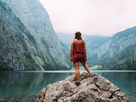 Woman in a red outfit stands on a rock by a clear alpine lake, facing misty cliffs and forested mountains.
