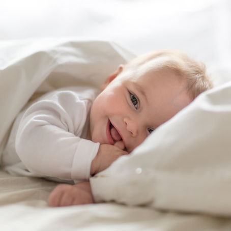 A smiling baby lies under a white blanket on a bed.