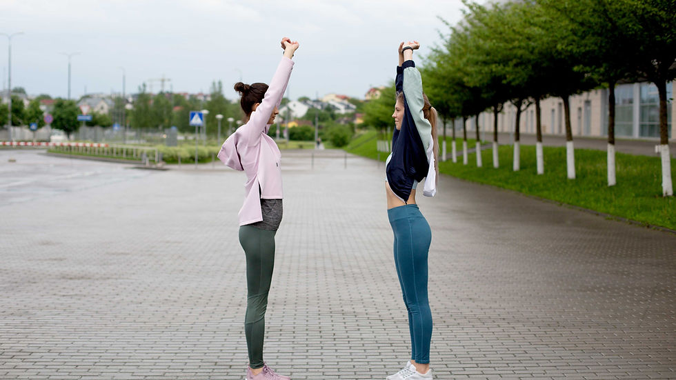 Hai phụ nữ mặc đồ tập đứng đối diện, giơ tay duỗi người trên vỉa hè ướt dưới trời âm u, hàng cây xanh bên đường.
