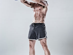 A muscular, shirtless man doing a kettlebell swing in a bright studio. Wears gray shorts, lime socks, and teal shoes. Intense focus.