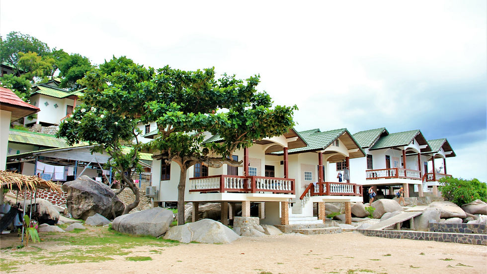 Houses with green roofs and balconies on a sandy beach, surrounded by trees and large rocks under a cloudy sky. People visible on balconies.