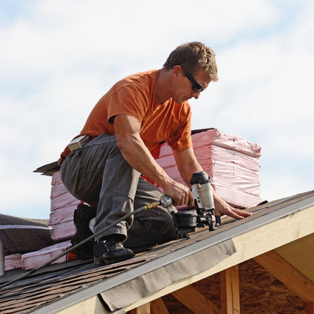 Roofer installing shingles on a residential home under clear sky with roofing materials stacked nearby