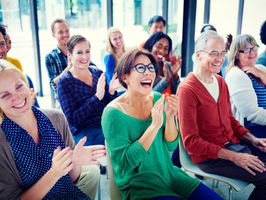 Diverse audience of adults smiling and applauding in a bright room with large windows. Engaged and joyful atmosphere, representing improved communication and social connection with hearing aids.