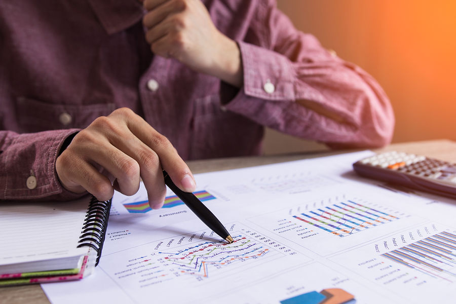 A person points at graphs and charts with a pen on a desk, surrounded by documents and a calculator, in warm lighting.