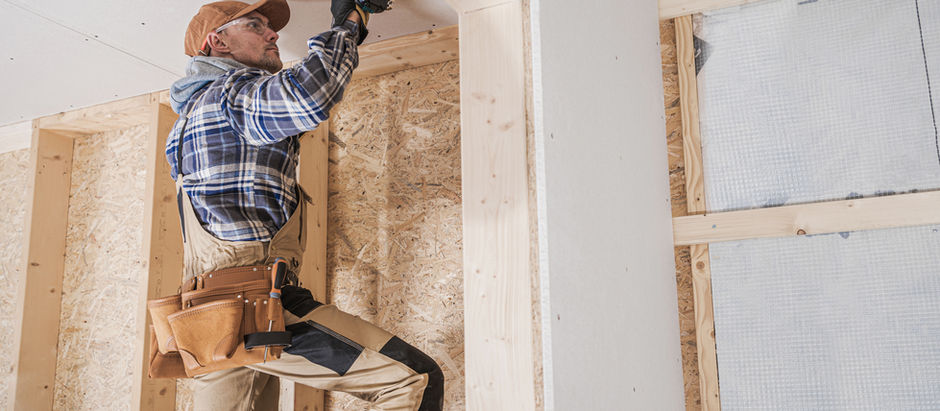 Worker in plaid shirt and cap uses a drill on a ceiling, standing on a ladder in a wooden-framed room, wearing a tool belt.