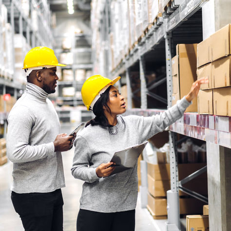 Two warehouse workers wearing yellow safety helmets conducting inventory checks with a clipboard and mobile device, surrounded by shelves stacked with cardboard boxes—representing logistics, supply chain management, and smart inventory solutions.