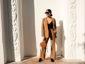 Woman wearing a camel tailored pantsuit and black top, holding a round brown handbag, standing against a white architectural wall — minimalist fall capsule wardrobe 2025 fashion.
