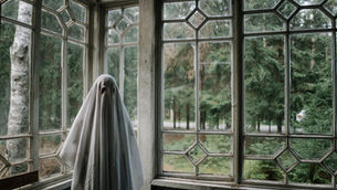 Ghostly figure in a white sheet stands in a rustic room with large, patterned windows overlooking a dense forest, creating a mysterious mood.