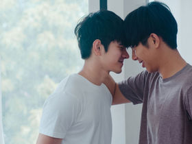 Two young Asian men stand forehead to forehead by a bright window, smiling softly in an intimate, affectionate moment.