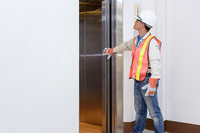 Side profile of a lift inspector wearing a white hard hat and safety glasses looking at a lift door