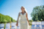A bride in a white gown and veil smiles joyfully while walking down an outdoor aisle