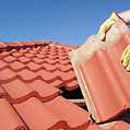 Roof repairs, worker with yellow gloves replacing red tiles or shingles on house with blue