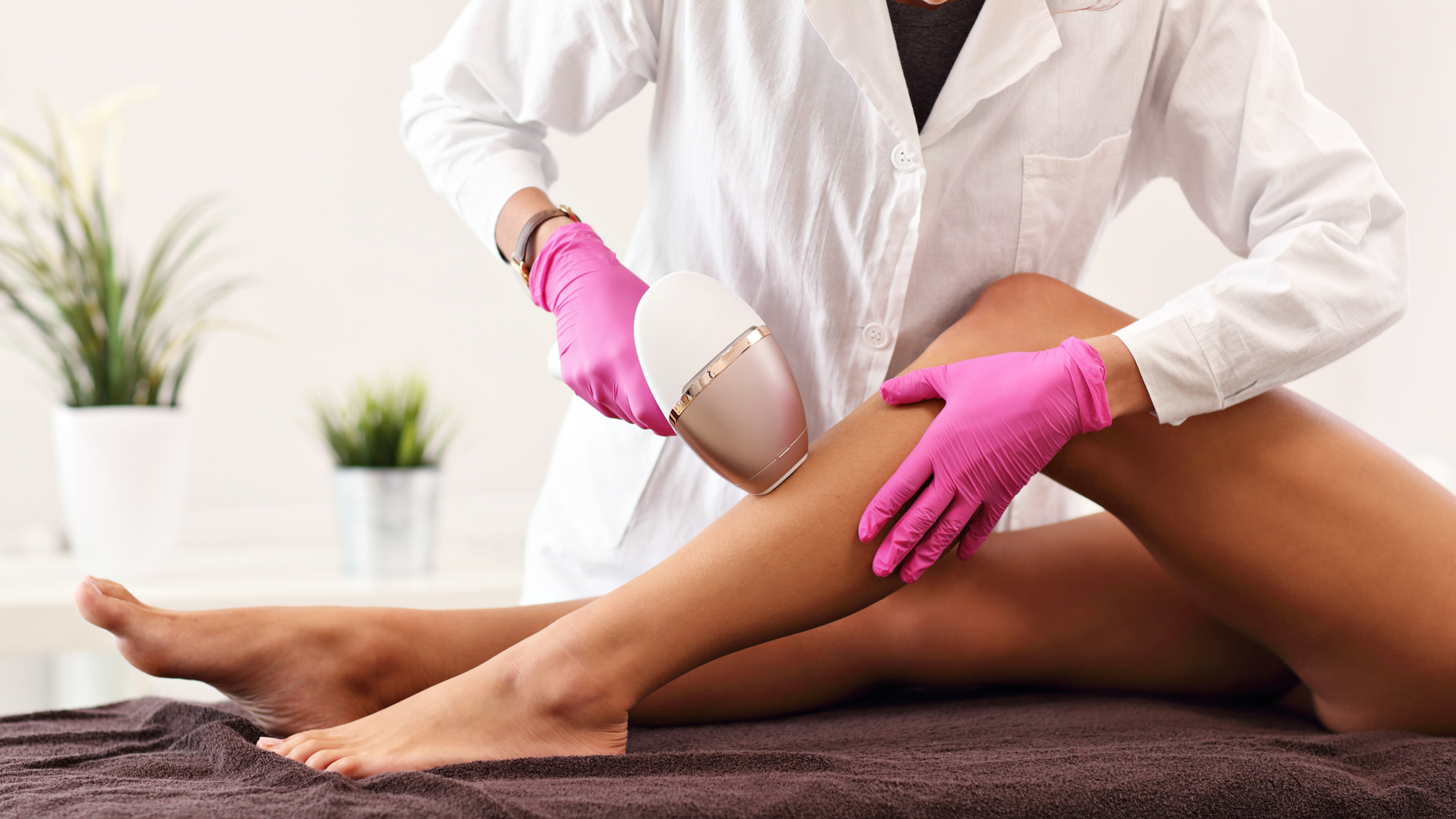 Esthéticienne en blouse blanche et gants roses épile la jambe d’une cliente avec un appareil, en cabine spa.